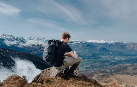 Un ami assis face à la vallée enneigée, portant un sac à dos Haize, admirant le paysage montagnard avec un air détendu et contemplatif.